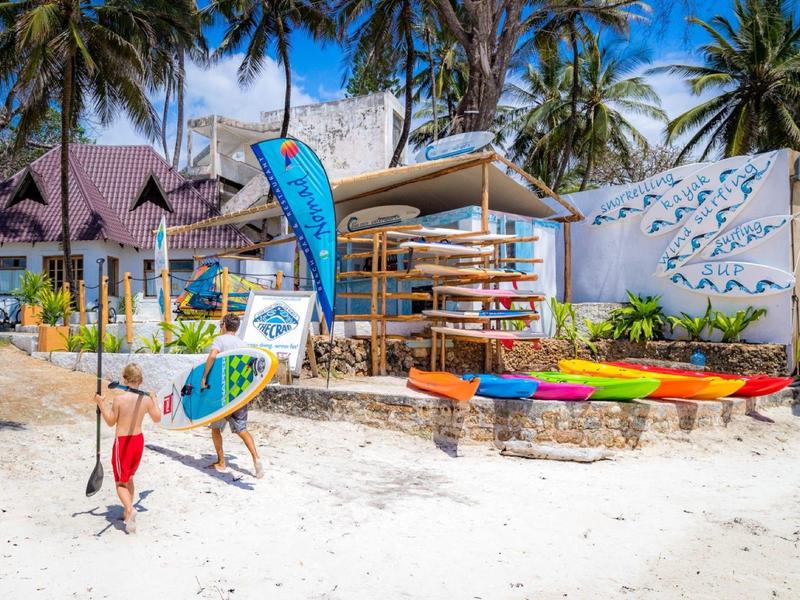 Colorful kayaks and surfboards on a sandy beach in front of a tropical surf rental with palm trees.