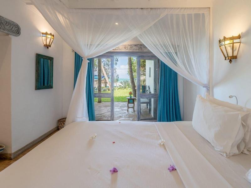 Bright hotel room with large bed, white curtains, and view of terrace and garden.
