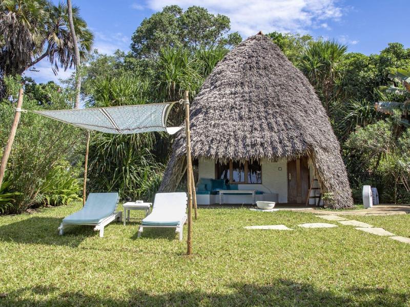 Small thatched vacation hut with two lounge chairs and sunshade in the garden.