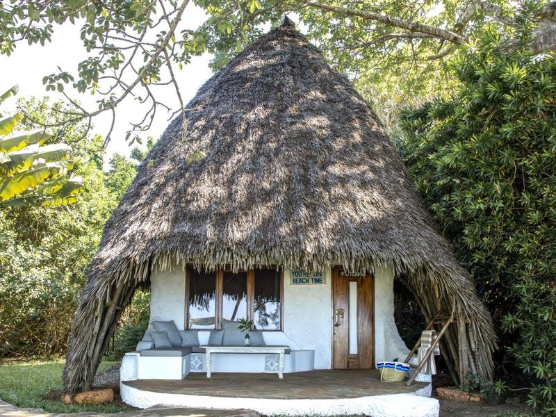 Small thatched hut with pointed roof and white veranda in a green setting.