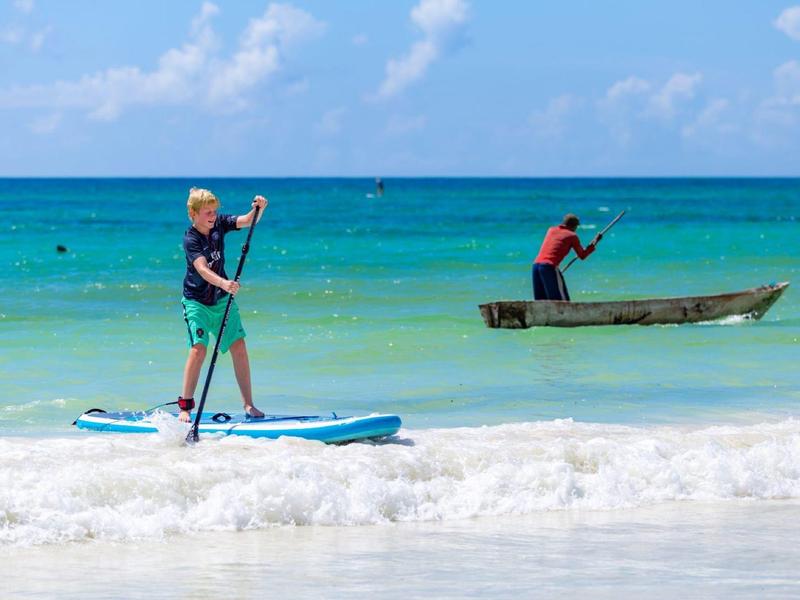 Person stands on a paddleboard in the sea while another rows a small boat.