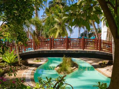 Pont en bois rouge au-dessus d'une piscine tropicale entourée d'une végétation luxuriante sous un ciel bleu.