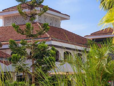 Bâtiment d'hôtel avec toit rouge entouré de palmiers et de hautes plantes sous un ciel bleu.