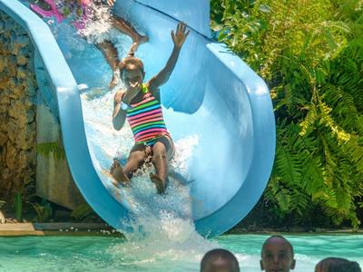 Des enfants glissent sur un toboggan bleu dans une piscine avec une végétation luxuriante en arrière-plan.
