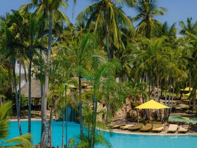 Une piscine tropicale avec des palmiers et des parasols jaunes sous un ciel bleu clair.