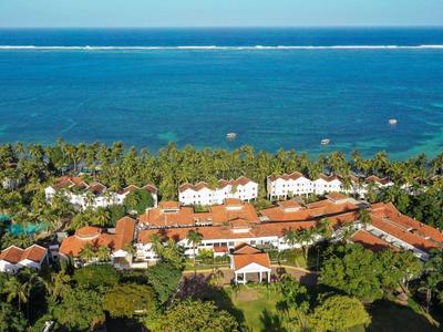 Vue aérienne d'un hôtel en bord de mer avec toits rouges, palmiers verts et mer bleue en arrière-plan.