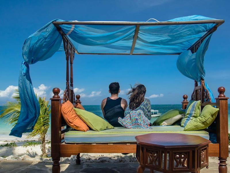 Couple se détend sur un lit à baldaquin sur la plage avec vue sur l'océan et ciel bleu.