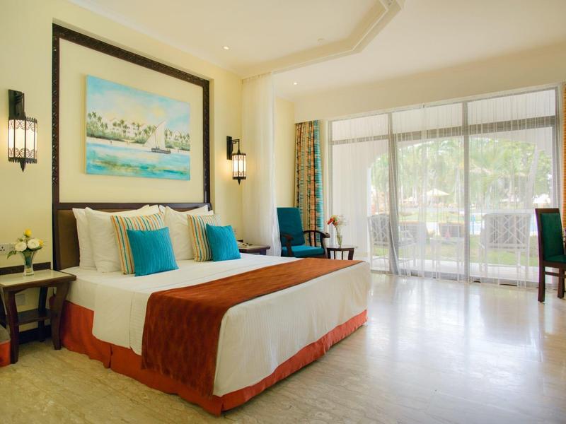 Chambre d'hôtel lumineuse avec lit double, coussins bleus et grande fenêtre donnant sur la terrasse.