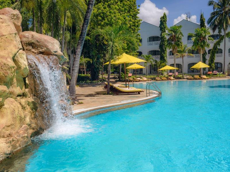 Piscine ensoleillée avec toboggan entourée de palmiers et de chaises longues dans une station tropicale.