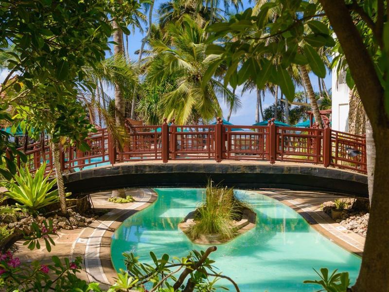 Pont en bois rouge au-dessus d'une piscine tropicale entourée d'une végétation luxuriante sous un ciel bleu.