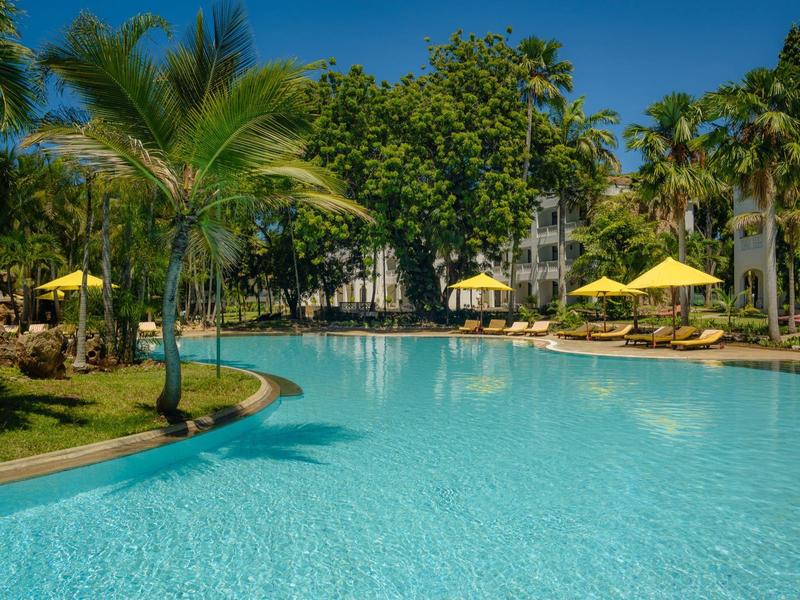 Grande piscine extérieure avec palmiers et parasols jaunes dans un jardin d'hôtel tropical.
