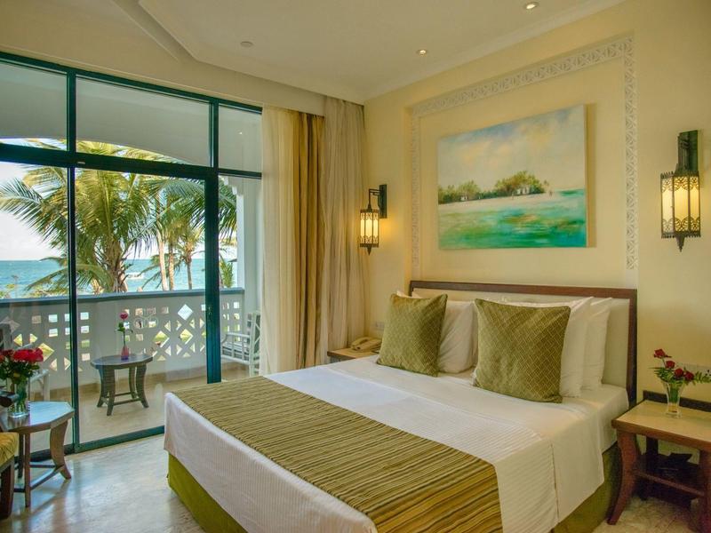 Chambre d'hôtel lumineuse avec grand lit, oreillers verts et vue sur la mer à travers de grandes baies vitrées.