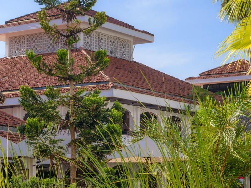 Bâtiment d'hôtel avec toit rouge entouré de palmiers et de hautes plantes sous un ciel bleu.