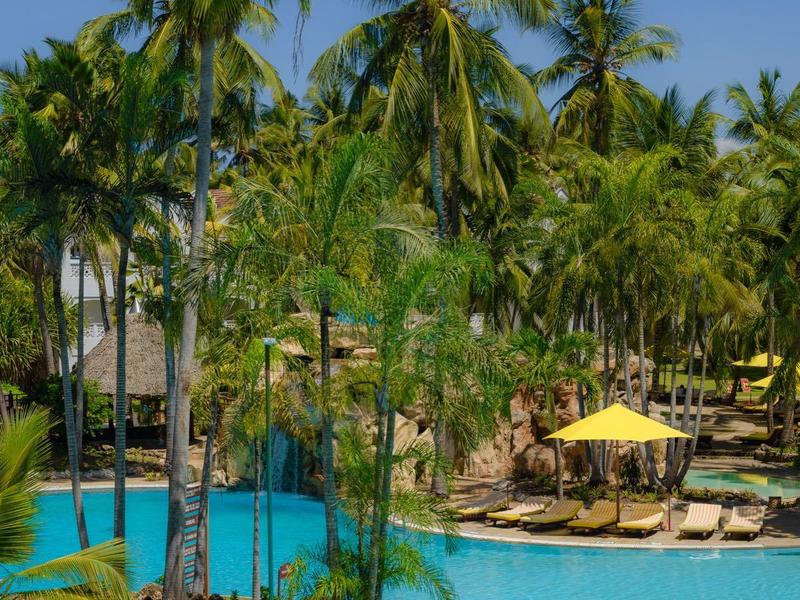 Une piscine tropicale avec des palmiers et des parasols jaunes sous un ciel bleu clair.