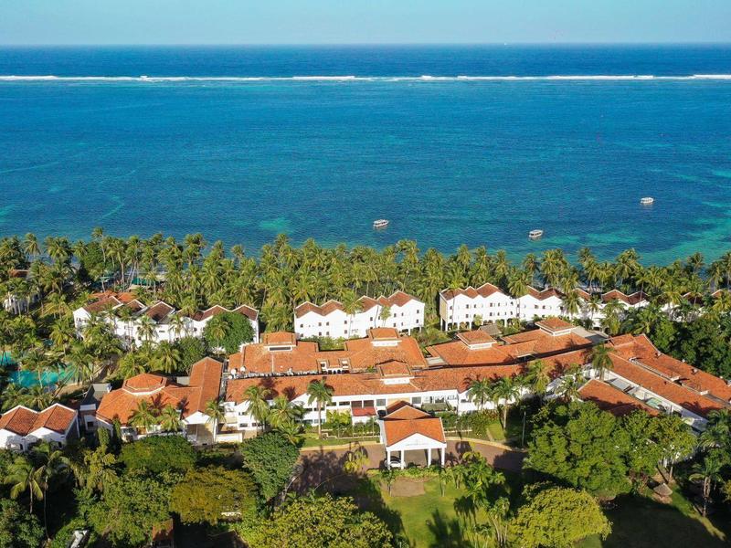 Vue aérienne d'un hôtel en bord de mer avec toits rouges, palmiers verts et mer bleue en arrière-plan.