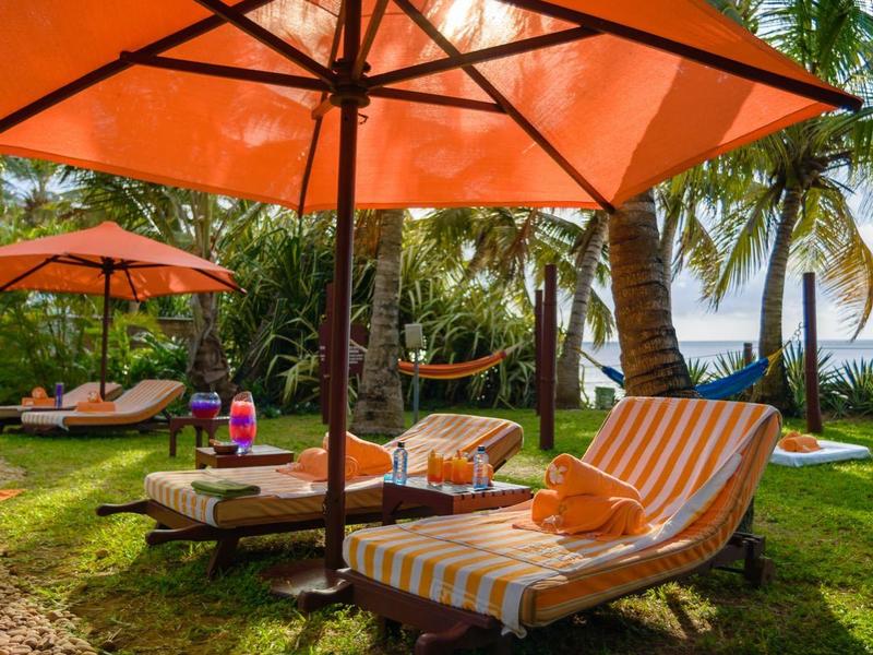 Deux chaises longues avec coussins rayés sous un grand parasol orange dans un jardin tropical.