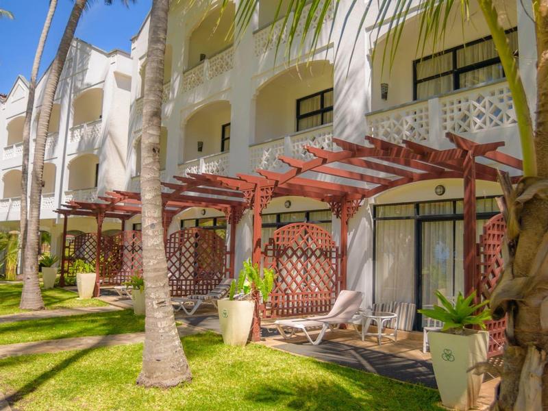 Terrasse d'hôtel avec chaises longues, palmiers et pelouse verte sous un ciel bleu.
