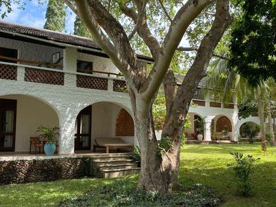 Grande maison blanche à deux étages avec des vérandas entourée d'arbres et de pelouse.