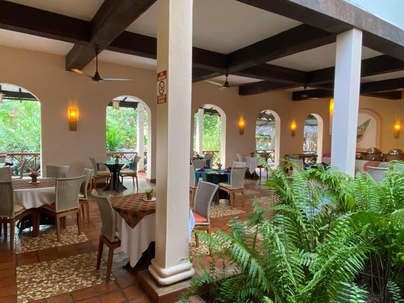 Ristorante luminoso con soffitto a travi di legno, tavoli apparecchiati e piante verdi.