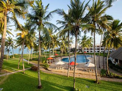 Hotelanlage mit Pool umgeben von Palmen und grüner Wiese unter blauem Himmel.
