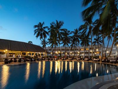 Abendaufnahme eines beleuchteten Hotelpools umgeben von Palmen und Liegestühlen.