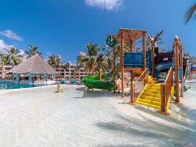 Kinder-Spielplatz mit Rutschen am sandigen Poolbereich eines Hotels unter blauem Himmel.