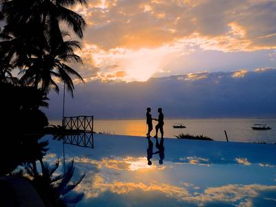 Zwei Personen stehen am Rand eines Pools mit Palmen, Sonnenuntergang im Hintergrund.