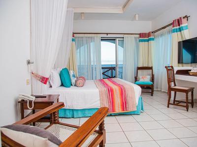 Světý hotelový pokoj s manželskou postelí, barevnými závěsy a výhledem na moře skrz velká okna.