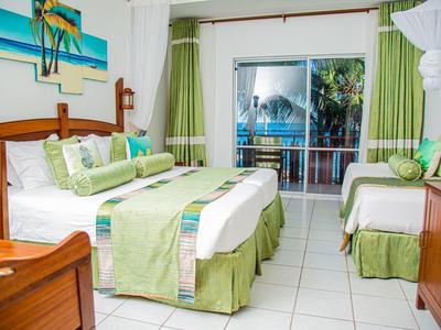 Světý hotelový pokoj se dvěma postelemi, zelenými závěsy a balkonem s výhledem na palmy.