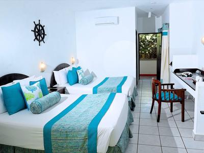 Světý hotelový pokoj se dvěma postelemi, námořnickými dekorativními polštáři a modro-zelenými akcenty.