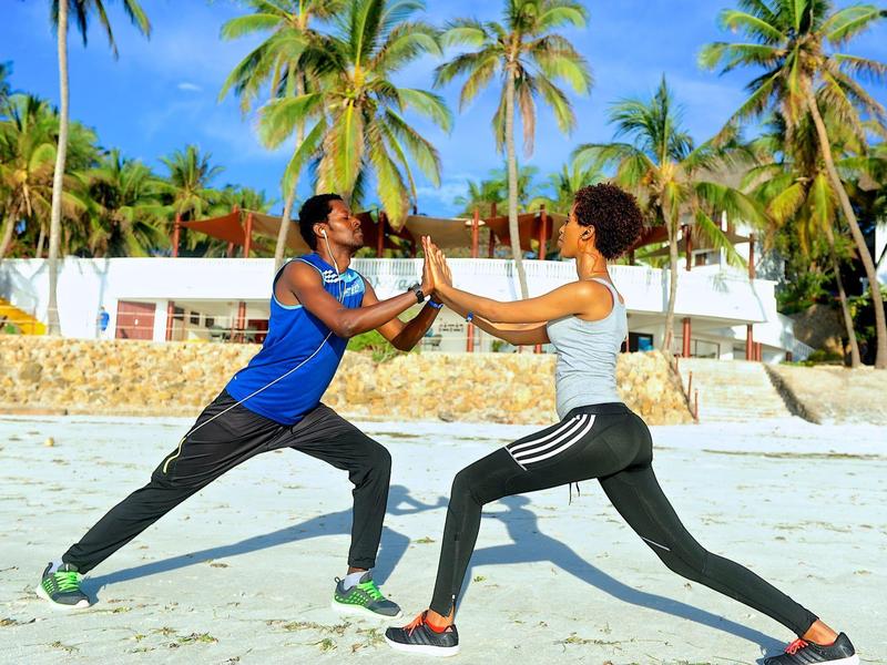 Dvě osoby cvičí jógu na pláži před hotelem s palmami.