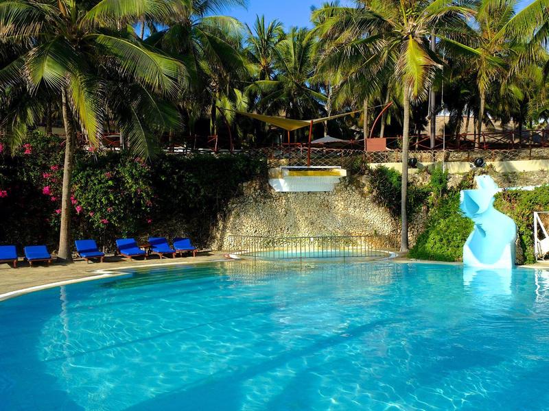 Modrý bazén s palmami, lehátky a tobogánem pod modrou oblohou.