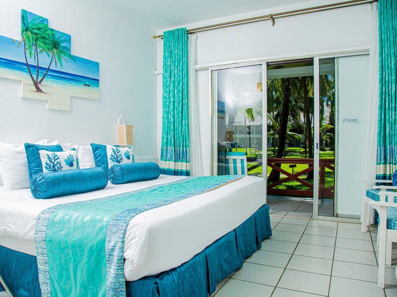 Světle zařízený hotelový pokoj s manželskou postelí a přístupem na balkon s výhledem na zeleň.