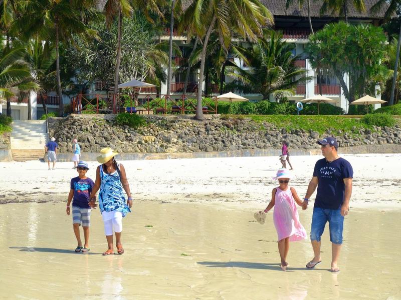 Pláž s lidmi chodícími blízko vody a tropickými rostlinami v pozadí.