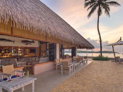 Strandrestaurant mit Strohdach, Holzmöbeln und Palmen am Wasser bei Sonnenuntergang.