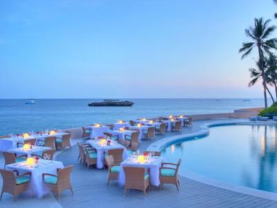 Restaurant-Tische mit Kerzen am Meer neben einem geschwungenen Pool und Palmen bei Abenddämmerung.