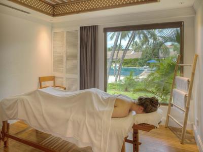 Helle Massagekabine mit Holzboden, Decke aus Holz, Liege und Blick auf Palmen und Wasser draußen.