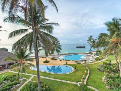 Blick auf tropischen Poolbereich mit Palmen, Rasenflächen, weißem Sandstrand und ruhigem Meer im Hintergrund.