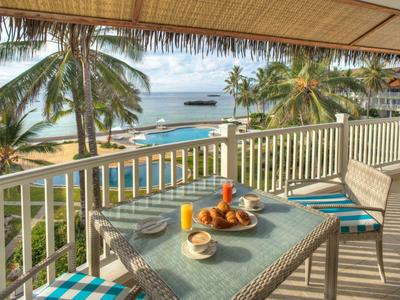 Balkon mit Tisch und Stühlen, tropische Palmen und Meer im Hintergrund, sonniger Strandblick.