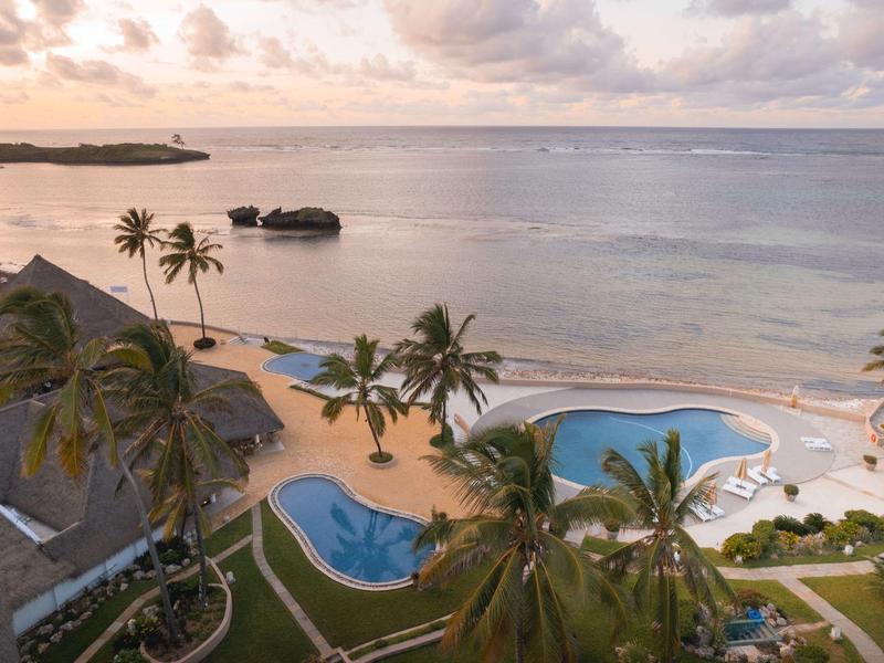Strand mit Palmen, zwei blauen Pools und ruhigem Meer unter wolkigem Himmel im Abendlicht.