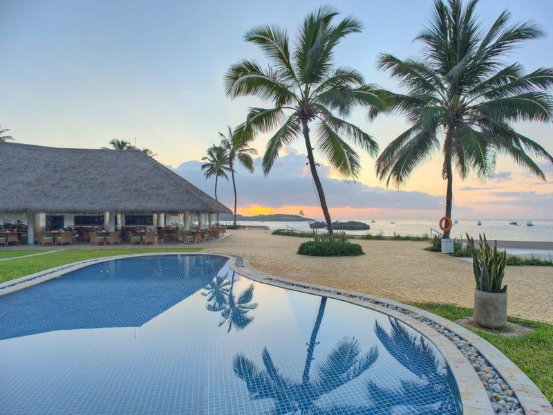 Ein Pool mit klarem Wasser, Palmen und Strand bei Sonnenuntergang mit ruhigem Meer im Hintergrund.