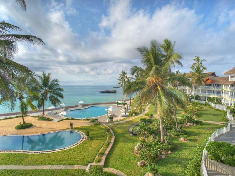 Strandhotel mit Palmen, zwei Pools, blauer Lagune und bewölktem Himmel am Meeresufer.