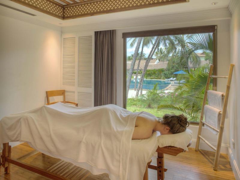 Helle Massagekabine mit Holzboden, Decke aus Holz, Liege und Blick auf Palmen und Wasser draußen.