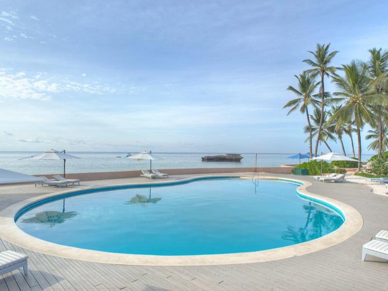 Runder Pool mit klarem Wasser, Liegen und Palmen am Meer unter blauem Himmel.