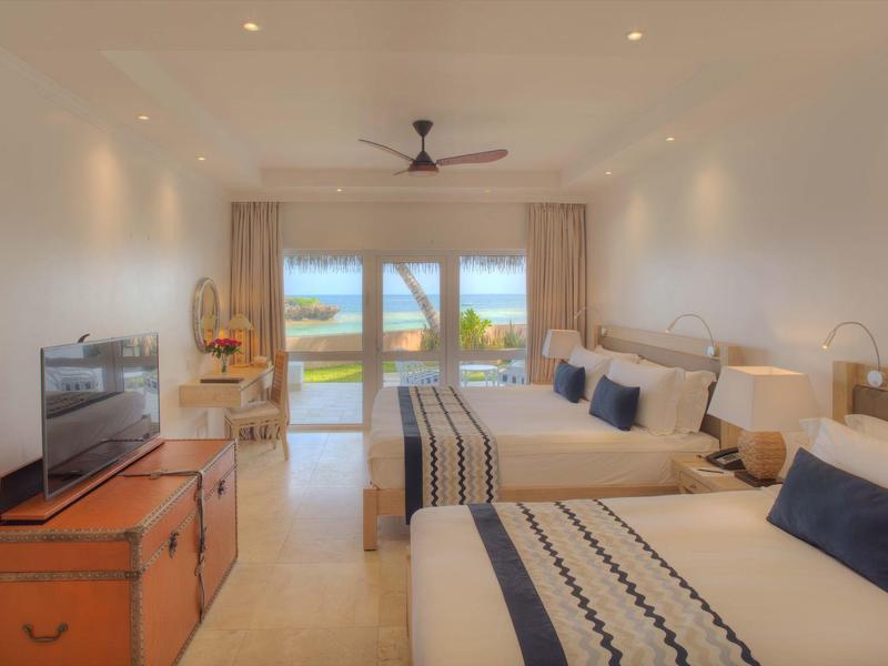 Helles Hotelzimmer mit zwei Queensize-Betten, Fernseher, Ventilator und Meerblick durch große Fenster.