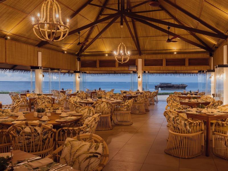 Offenes Restaurant mit Holzdach, runden Tischen und Stühlen, warmem Licht und Meerblick bei Dämmerung.