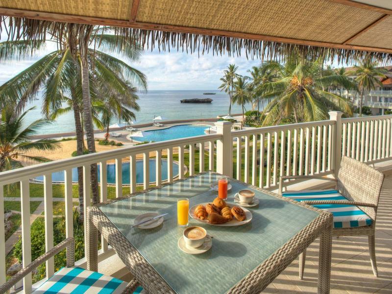 Balkon mit Tisch und Stühlen, tropische Palmen und Meer im Hintergrund, sonniger Strandblick.