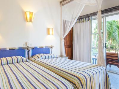 Licht ingerichte slaapkamer met twee bedden en uitzicht op een terras met palmbomen.