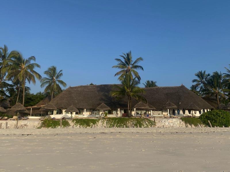 Strandresort met rieten daken, palmbomen en parasols op zandstrand bij heldere lucht.