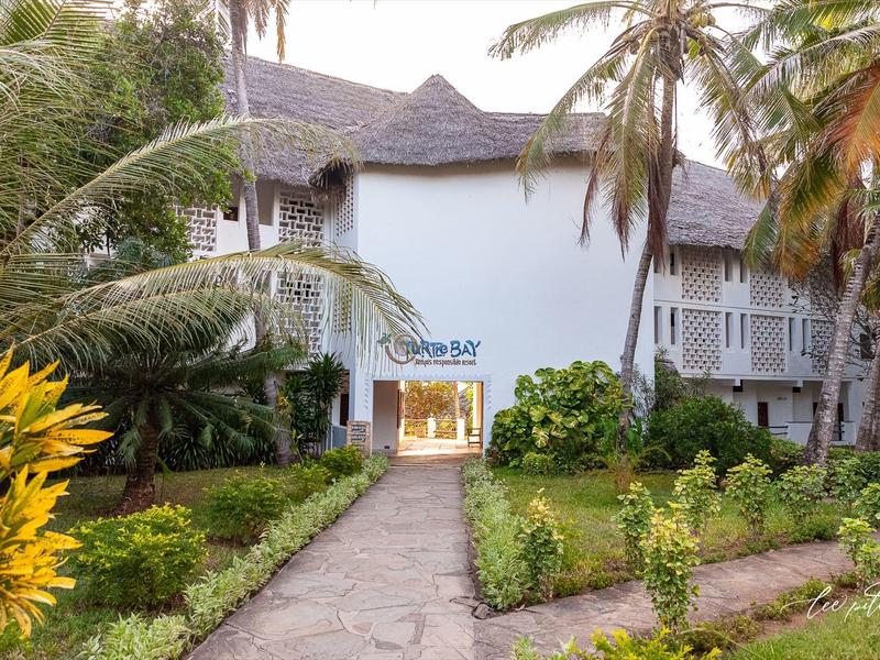 Wit hotelgebouw met puntdaken, omgeven door palmbomen en een verzorgde tuin.